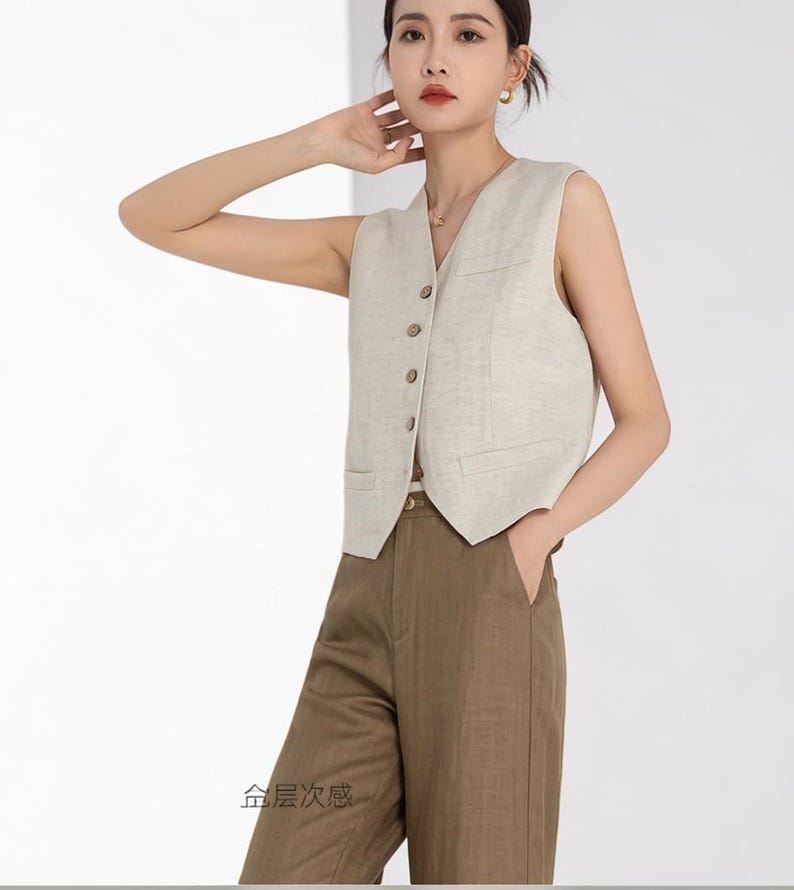 May include: A beige sleeveless vest with a V-neck, button closure, and two front pockets. The vest is paired with brown trousers. The outfit is made of a light-colored fabric, suggesting a casual or business casual style.