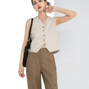May include: A beige sleeveless vest with a V-neck and wooden buttons. The vest is paired with brown trousers. A black shoulder bag hangs on the left side. The outfit is a neutral color palette.
