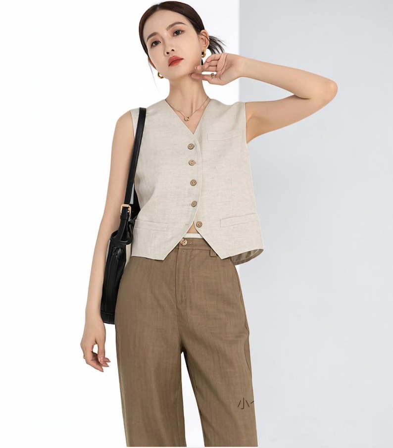 May include: A beige sleeveless vest with a V-neck and wooden buttons. The vest is paired with brown trousers. A black shoulder bag hangs on the left side. The outfit is a neutral color palette.