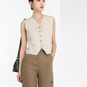 May include: A beige linen vest with a V-neck and button closure, paired with brown trousers. The vest has two small pockets and is sleeveless. The trousers have a straight leg design. The outfit is a neutral color.