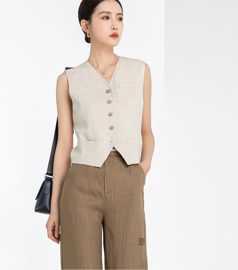 May include: A beige linen vest with a V-neck and button closure, paired with brown trousers. The vest has two small pockets and is sleeveless. The trousers have a straight leg design. The outfit is a neutral color.