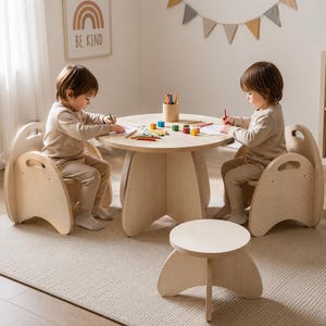 Puede incluir: Un conjunto de muebles infantiles de madera clara que consta de una mesa redonda, dos sillas con asas recortadas y un pequeño taburete circular. La superficie de la mesa contiene un portalápices de madera con lápices de colores y bloques.