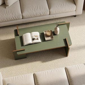 May include: A rectangular sage green coffee table with a flat top and interlocking wooden side supports. The table surface holds an open book, a small rectangular tray containing a glass, and a smooth, light-colored ceramic vase.