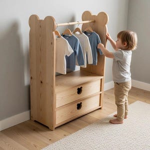 May include: A light-coloured wooden clothing rack featuring bear-shaped side panels and a horizontal hanging rod. The unit includes two drawers with bear-shaped cutouts for handles, holding four long-sleeved shirts in white and blue tones.