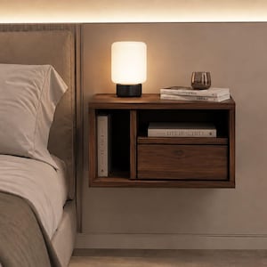 Op de afbeelding: Een aan de muur gemonteerd zwevend nachtkastje van donker hout met een open verticaal schap aan de linkerzijde en een lade aan de rechterzijde. Bovenop staat een cilindervormige witte lamp met een zwarte voet, een glas en twee boeken.
