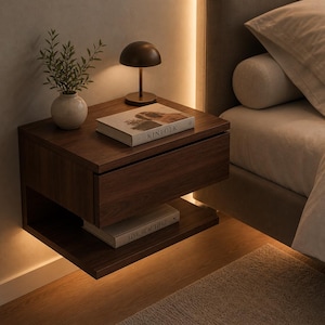 May include: A floating dark wood nightstand with a drawer and a lower shelf. A small lamp and a vase with greenery sit on top. The book "KINFOLK" is visible on the top. A second book, "LIVE BEAUTIFUL", is on the shelf below.