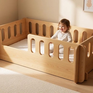 May include: A light-coloured wooden toddler bed featuring rounded vertical slats on all sides, a low-profile frame, and a white mattress. The bed is designed with a small opening on one side and has a smooth, natural wood grain finish throughout the structure.