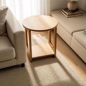 Puede incluir: Una mesa auxiliar redonda de madera con un acabado natural claro, que presenta una parte superior circular y un estante inferior cuadrado sostenido por cuatro patas verticales. La mesa tiene un patrón de veta de madera visible y abierta.