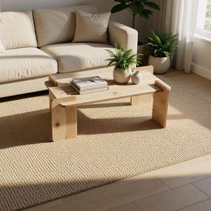 May include: A light-colored wooden coffee table with a rectangular top and two side supports, featuring a stack of books and two small white ceramic vases with green plants on its surface, positioned in a room with a beige sofa and a textured rug.