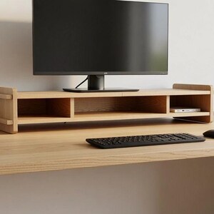 Op de afbeelding: Een monitorstandaard van lichtgekleurd hout met drie open opbergvakken, geplaatst op een bijpassend houten bureau. Een zwarte computermonitor staat op de standaard, terwijl een zwart toetsenbord en een zwarte computermuis op het bureau liggen.