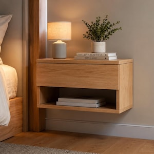 May include: A wall-mounted oak bedside table with a drawer and shelf. A small lamp, a plant in a pot, and books are on top. The table is next to a bed, against a beige wall.