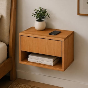 May include: A floating wooden nightstand with a single drawer and an open shelf. A small potted plant and a smartphone sit on top. Two books are visible on the shelf. The nightstand is made of light-colored wood.