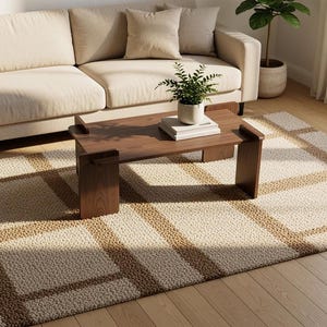 May include: A rectangular area rug featuring a textured, cream-colored base with a prominent grid pattern formed by thick, brown, woven lines. The rug is positioned on a light-toned wooden floor, partially underneath a dark brown wooden coffee table.