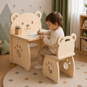 Op de afbeelding: Een lichtgekleurde houten kinderbureau en stoelset. Het bureau heeft een berenvormige rug en pootvormige uitsparingen. Een kind zit aan het bureau en tekent met een groene potlood. Een kleine houten potlodenhouder staat op het bureau.