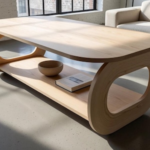 Puede incluir: Una mesa de centro de madera clara con una superficie rectangular de bordes redondeados y un estante inferior sostenido por dos grandes paneles laterales ovalados. Un cuenco peque&ntilde;o y sencillo y un libro cerrado descansan en el estante.