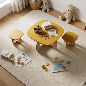 Puede incluir: Una mesa cuadrada de color amarillo mostaza con esquinas redondeadas y dos taburetes a juego, cada uno con patas de madera clara en forma de cruz. La mesa contiene dos recipientes con lápices de colores, libros y un dibujo colorido.