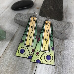 May include: A pair of green, yellow, and purple geometric earrings with a floral design. The earrings are made of wood and have silver hooks.