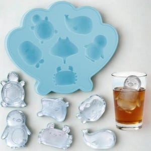 May include: A light blue silicone ice cube tray in a seashell shape, filled with various sea creature molds. Includes ice cubes shaped like a turtle, penguin, crab, and whale. A glass of iced tea contains a penguin ice cube.