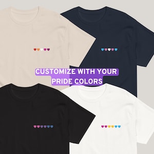 Puede incluir: Cuatro camisetas en crema, azul marino, negro y blanco, cada una con una fila de corazones bordados en diferentes colores de la bandera del orgullo. El texto "CUSTOMIZE WITH YOUR PRIDE COLORS" está superpuesto en la imagen.