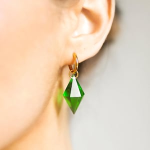 May include: A close-up of an ear with a gold hoop earring. The earring features a green, diamond-shaped charm. The charm is a faceted, emerald green colour, reflecting light. The earring is a simple, elegant design.