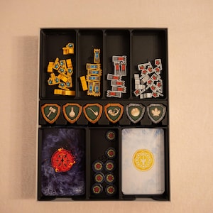Può includere: Un vassoio organizzatore in plastica nera per giochi da tavolo contenente vari componenti di gioco. Il vassoio ospita gettoni torre gialli e rossi, sei gettoni a forma di scudo con icone, un mazzo di carte blu scuro, un mazzo di carte bianche e gettoni circolari.