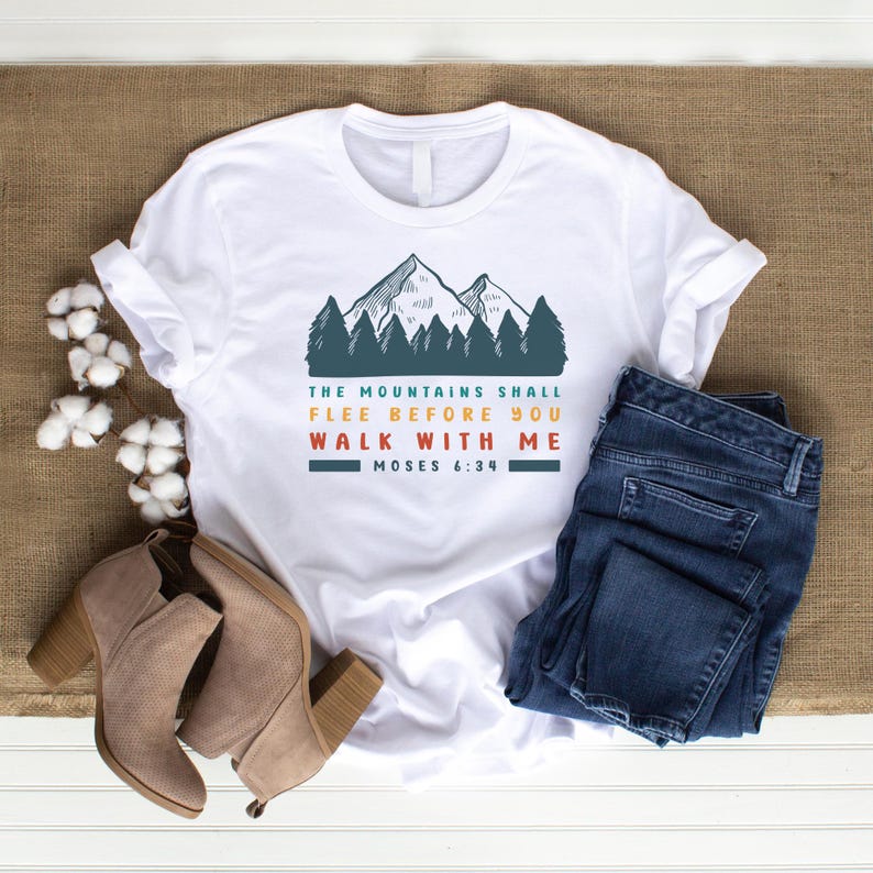May include: White t-shirt with a mountain and forest graphic in teal and orange, and the text "THE MOUNTAINS SHALL FLEE BEFORE YOU WALK WITH ME MOSES 6:34". The shirt is paired with blue jeans and tan ankle boots.