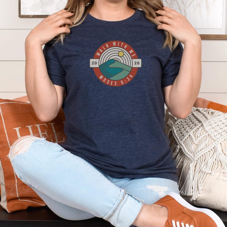 May include: Navy blue t-shirt featuring a circular graphic with the text "WALK WITH ME" and "MOSES 6:34". The graphic includes a mountain and sun design in shades of blue, green, and yellow. The shirt is a casual wear item.