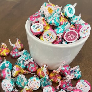 Pode incluir: Uma tigela branca cheia de beijos de chocolate embrulhados em papel alumínio colorido com designs de Páscoa. Os designs incluem coelhos, ovos e as palavras "Feliz Páscoa", "Beijos de coelho" e "Abraços".