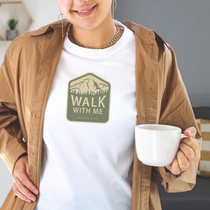 May include: White t-shirt with a green and tan mountain graphic that reads "WALK WITH ME" and "MOSES 6:34". The shirt is worn with a brown jacket. A white mug is held in the hand.