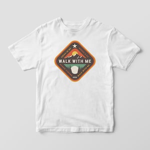 May include: White t-shirt featuring a diamond-shaped graphic with the text "WALK WITH ME". The graphic includes a mountain scene with a sun, and the phrases "ABIDE IN ME", "STAND STRONG", and "MOSES 5:24".