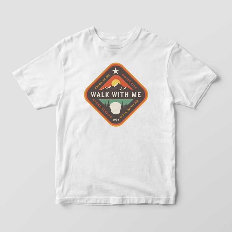 May include: White t-shirt featuring a diamond-shaped graphic with the text "WALK WITH ME". The graphic includes a mountain scene with a sun, and the phrases "ABIDE IN ME", "STAND STRONG", and "MOSES 5:24".