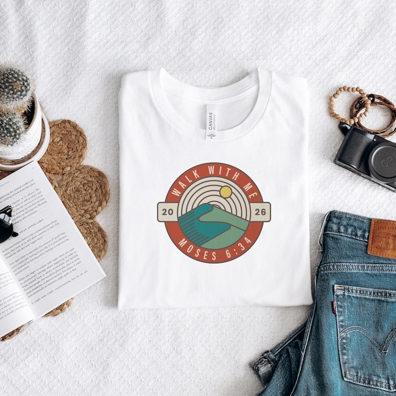 May include: A white t-shirt featuring a circular graphic with the text "Walk With Me" and "Moses 6:34". The design includes a mountain scene with a yellow sun, teal and brown colors. The shirt is laid flat on a white surface.