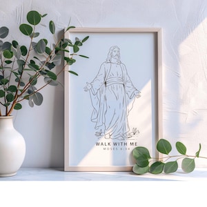 May include: A framed print featuring a line drawing of a figure with outstretched arms. The artwork is in black and white, with the text "WALK WITH ME MOSES 6:34" at the bottom. The frame is light-colored wood. A vase with greenery is to the left.