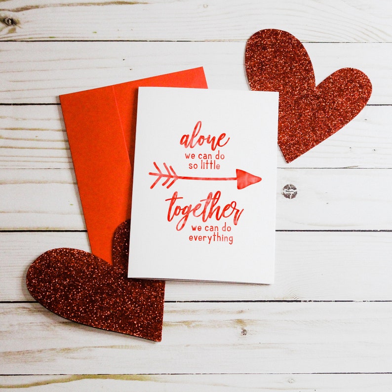Puede incluir: Una tarjeta blanca con letras rojas que dice "alone we can do so little together we can do everything". La tarjeta tiene una flecha roja apuntando hacia la derecha. La tarjeta est&aacute; rodeada de dos corazones rojos de purpurina.