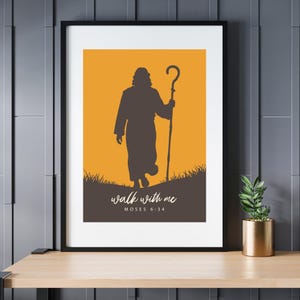 May include: Framed art print featuring a silhouette of a figure holding a staff, set against an orange background. The text "walk with me" and "MOSES 6:34" are displayed in white. The artwork is framed in black and rests on a wooden surface.