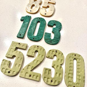 May include: A collection of decorative house numbers in various colors. The numbers include 85 in cream, 103 in teal, 5230 in olive green. Each number has a unique textured design and pre-drilled holes for mounting.