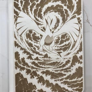 May include: A rectangular white binder featuring a brown monochromatic illustration of the legendary Pokémon Lugia flying above a turbulent ocean with stylised waves and rocky formations. The cover has a textured finish and a stitched border edge.