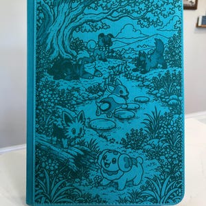Puede incluir: Una cubierta de cuaderno rectangular de color verde azulado con una ilustración detallada en verde azulado oscuro de varias criaturas pequeñas en un entorno forestal. La escena incluye árboles, flores y un arroyo con piedras.