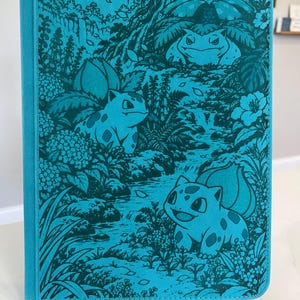 May include: A rectangular turquoise notebook featuring a dark teal monochromatic illustration of three creatures resembling Bulbasaur, Ivysaur, and Venusaur nestled within a dense jungle landscape with waterfalls, ferns, and various tropical flowers.