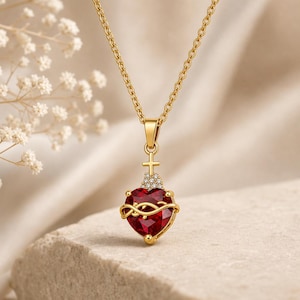 Puede incluir: Un collar de tono dorado con un colgante de piedra roja en forma de corazón. El colgante está rodeado por una corona de espinas de alambre dorado, rematado con una pequeña cruz dorada y un grupo de piedras blancas, colgado de una cadena fina.