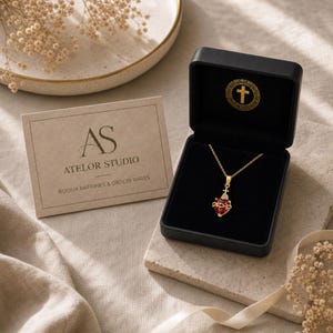 Peut inclure: Un collier doré avec un pendentif en forme de cœur rouge orné d'une petite croix et d'une couronne d'épines, présenté dans un coffret noir avec un logo croix doré. Une carte beige indique AS ATELOR STUDIO BIJOUX RAFFINÉS & OBJETS RARES.