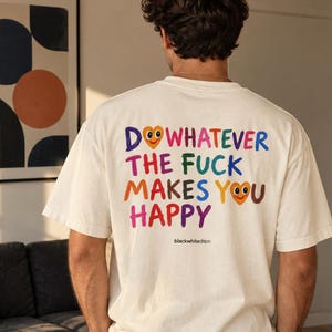 Op de afbeelding: Een off-white t-shirt met korte mouwen en de kleurrijke tekst DO WHATEVER THE FUCK MAKES YOU HAPPY op de achterkant. De letters zijn in verschillende kleuren, met twee hartvormige smileys die de letter O vervangen. De tekst blackwhitedition staat eronder.