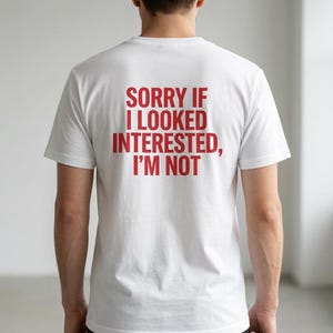 Peut inclure: Un t-shirt blanc à manches courtes avec un texte rouge en lettres capitales imprimé au dos qui indique, SORRY IF I LOOKED INTERESTED, I'M NOT. Le vêtement présente une encolure ronde et une coupe classique, mettant le texte en évidence.