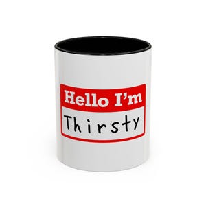 May include: A white ceramic mug with a black interior and rim. The front features a red rectangular name tag graphic with the white text Hello I'm and the word Thirsty written in black handwritten-style font centred below the red section.