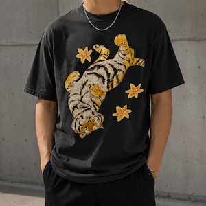 Può includere: Una t-shirt nera a maniche corte con una grande stampa grafica di una tigre sdraiata sulla schiena con le zampe in aria. La tigre ha strisce arancioni e nere ed è circondata da tre piccoli fiori gialli a cinque petali.