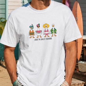 Peut inclure: Un t-shirt blanc à manches courtes orné d'un motif représentant quatre oies de dessin animé habillées pour la plage, dont une avec un tuba, une avec une tranche de pastèque, une en bikini et une avec une chemise à fleurs, avec le texte JUST A SILLY GOOSE.
