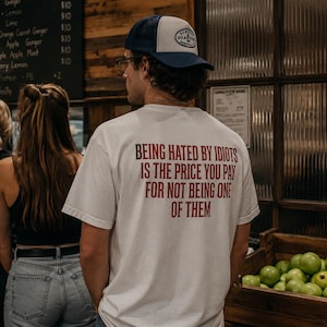 Peut inclure: Un t-shirt blanc à manches courtes avec un texte rouge au dos qui dit : BEING HATED BY IDIOTS IS THE PRICE YOU PAY FOR NOT BEING ONE OF THEM. Le t-shirt est porté avec une casquette de camionneur bleue et blanche avec un logo circulaire.