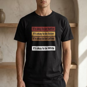 May include: Black crew neck t-shirt featuring the text "It's okay to be Native, Asian, Latino, White" in a stacked format. The text is in red, yellow, brown, and white. A simple, bold design.
