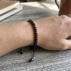May include: A bracelet featuring alternating dark brown wooden cylindrical beads and small metallic gray spherical beads strung on a black cord. The bracelet includes an adjustable macrame closure with two dangling black cords tipped with metallic gray beads.