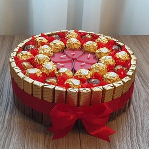 Könnte beinhalten: Eine runde Geschenkbox mit rotem Band und Schleife. Die Box ist mit Schokoriegeln an den Seiten verziert und mit goldverpackten Pralinen und herzförmigen Kerzen gefüllt. Auf einer Holzoberfläche platziert.